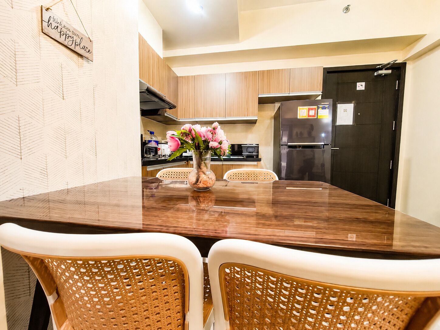 A dining area with a wooden table and two wicker chairs, a vase with flowers, and a kitchen in the background.