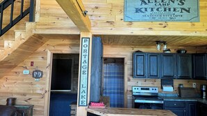 A rustic cabin interior with a wooden staircase, a kitchen area, and a sign reading "Allen's Camp Kitchen." - Charming 2-bedroom cabin in Portage Lake (Portage Lake)