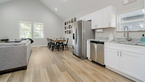 A modern kitchen with white cabinets, a stainless steel refrigerator, and a dishwasher. There is a dining area with a table and chairs. - BRAND NEW Spacious and Uniquely Musical Home near Downtown Indianapolis! (Indianapolis)