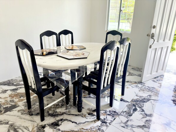 Une table de salle à manger ronde, accompagnée de six chaises, chacune dotée d’une structure noire et de coussins d’assise blancs. Le plateau de la table est en marbre blanc et elle est entourée d’un mur blanc.