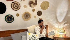 A person sitting on a bed with a lamp and a book on a bedside table.