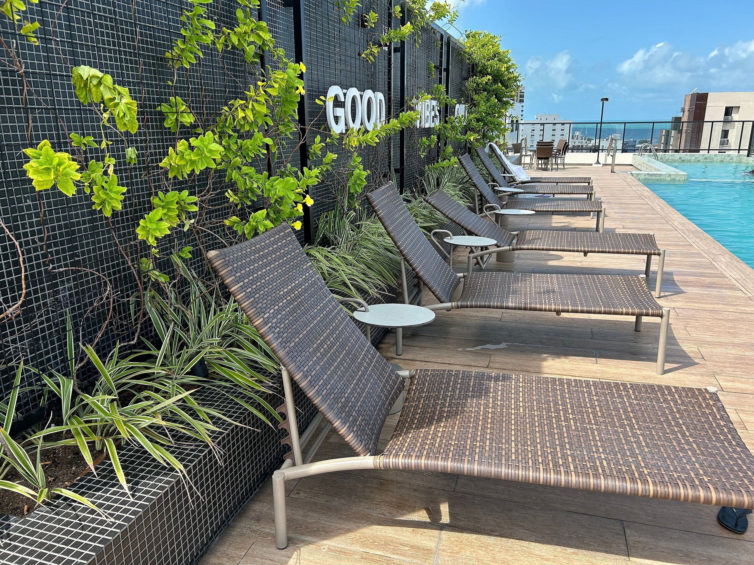 Uma área à beira da piscina com espreguiçadeiras e mesas, uma placa com a inscrição 'GOOD VIBES' e vista para a piscina.