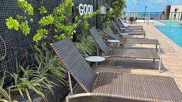 Uma área à beira da piscina com espreguiçadeiras e mesas, uma placa com a inscrição 'GOOD VIBES' e vista para a piscina.
