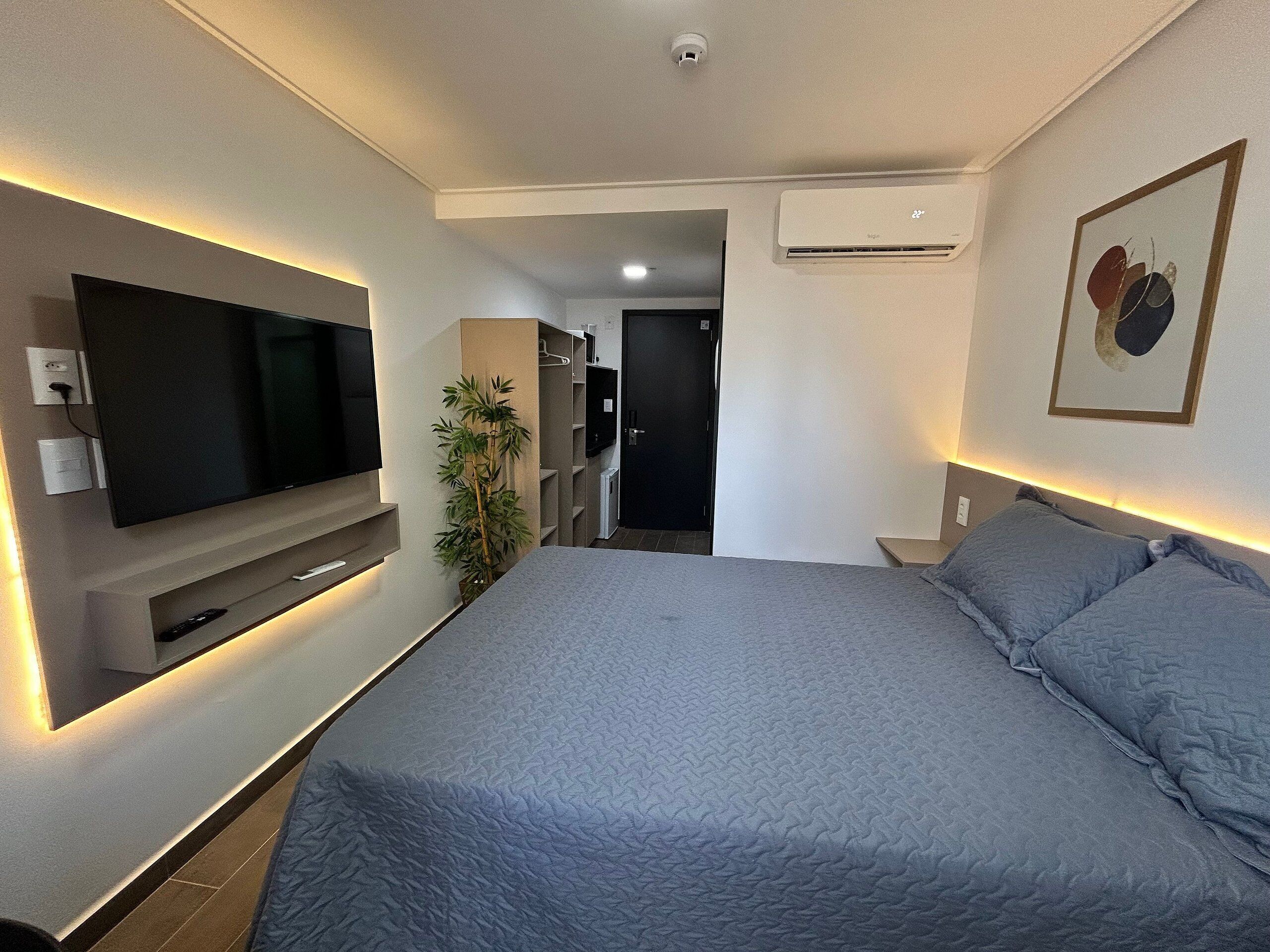 Quarto de hotel moderno com uma cama grande, uma televisão de tela plana na parede, uma planta em vaso e um aparelho de ar condicionado.
