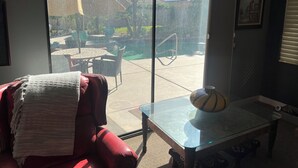 A room with a red leather chair, a glass table with a decorative vase, and a view of a backyard with a pool, palm trees, and a thatched-roof structure.