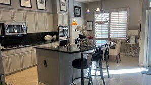 A modern kitchen with white cabinetry, a central island with bar stools, and a dining area with a view of the outdoors.