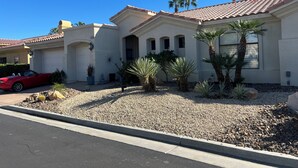 A two-story house with a red car parked in the driveway, palm trees, and a gravel front yard.