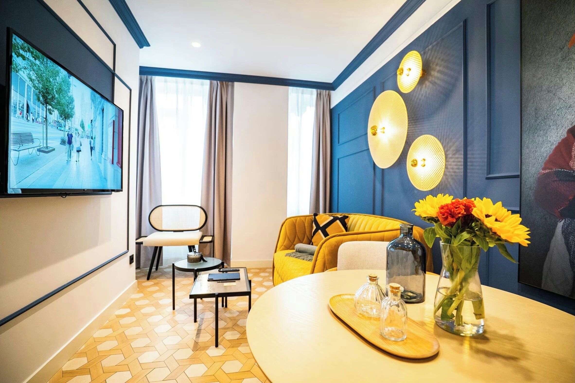 Habitación de hotel con televisor grande, sofá, mesa de comedor con un jarrón de flores y una lámpara fijada a la pared.
