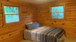 Ein Holzblockraum mit zwei Fenstern, einem Bett mit gestreifter Bettwäsche und einer Deckenleuchte.