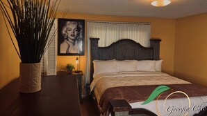 Een slaapkamer met een bed, nachtkastje, lamp, potplant en een ingelijst foto aan de muur.