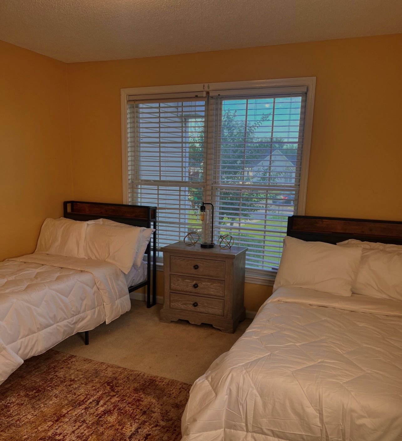 A bedroom with two single beds, a nightstand with a lamp, and a window with blinds.