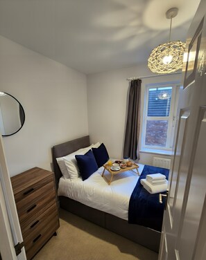 A neatly made bed with a tray of breakfast items, a bedside table, a wooden nightstand, and a window with curtains. - Dragonfly Abodes 4-Bed Sleeps 9 for teams in Bristol Near MoD + Parking (Stoke Gifford)