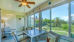 Pátio ensolarado com mesa, cadeiras, janelas amplas e ventilador de teto.