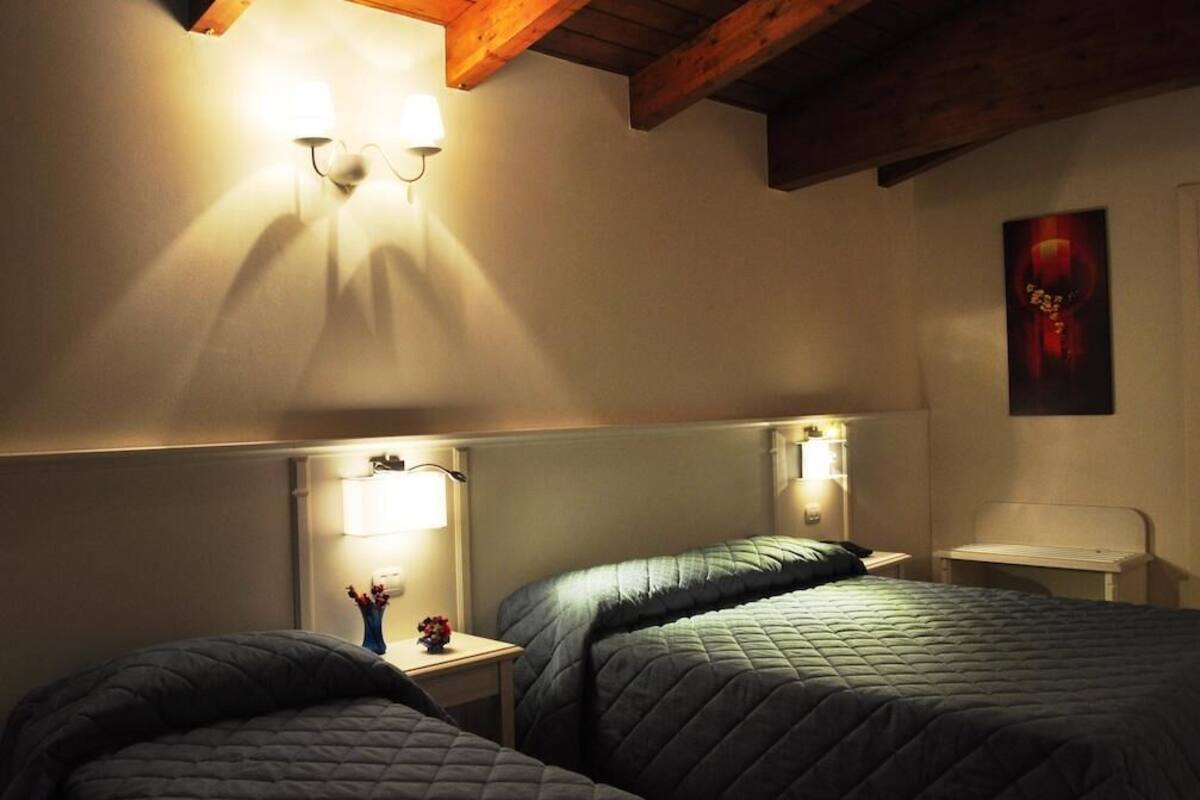 A hotel room with two beds, a nightstand with a lamp, a small table with a vase of flowers, and a painting on the wall.