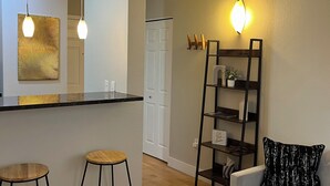 A modern living room with a white sofa, two wooden stools, a glass coffee table, a black metal shelf, and a wall-mounted light fixture. - The Lofty Loft - True City Living in Downtown Denver (Denver)