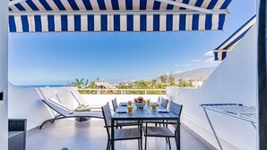 Una terraza en la azotea con una mesa preparada para dos, un toldo a rayas y vistas al mar y a las colinas.