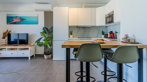 Cocina moderna con armarios blancos, encimera de madera y horno empotrado. Hay un televisor sobre un pedestal blanco, una planta en maceta y un cuadro con una escena costera en la pared.