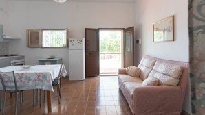 Une cuisine avec une table à manger, des chaises et un canapé. On y trouve un réfrigérateur, une fenêtre avec des barreaux et une porte donnant sur l’extérieur.