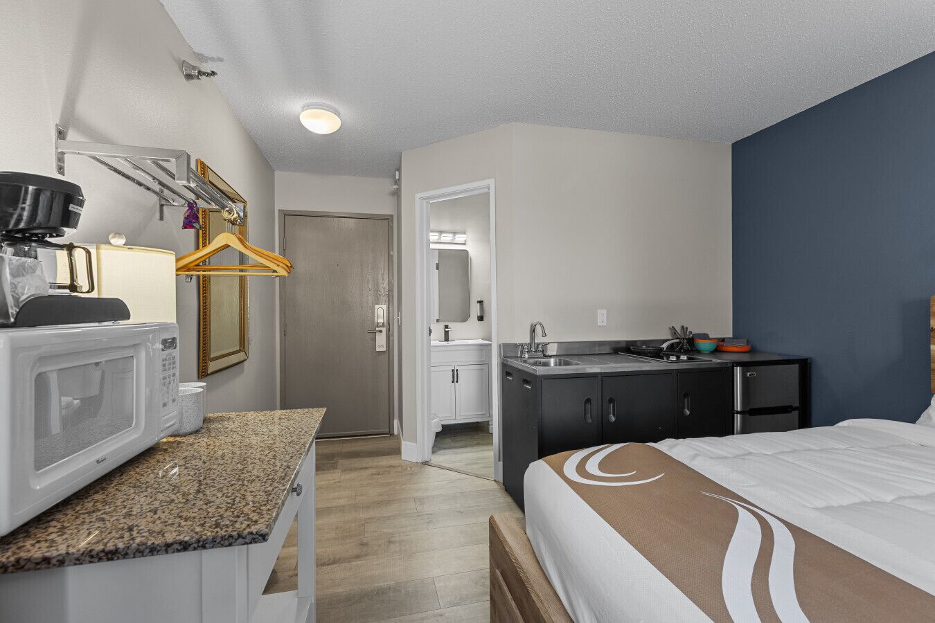 A compact hotel room with a kitchenette, a bed with a brown headboard, and a door leading to a bathroom.