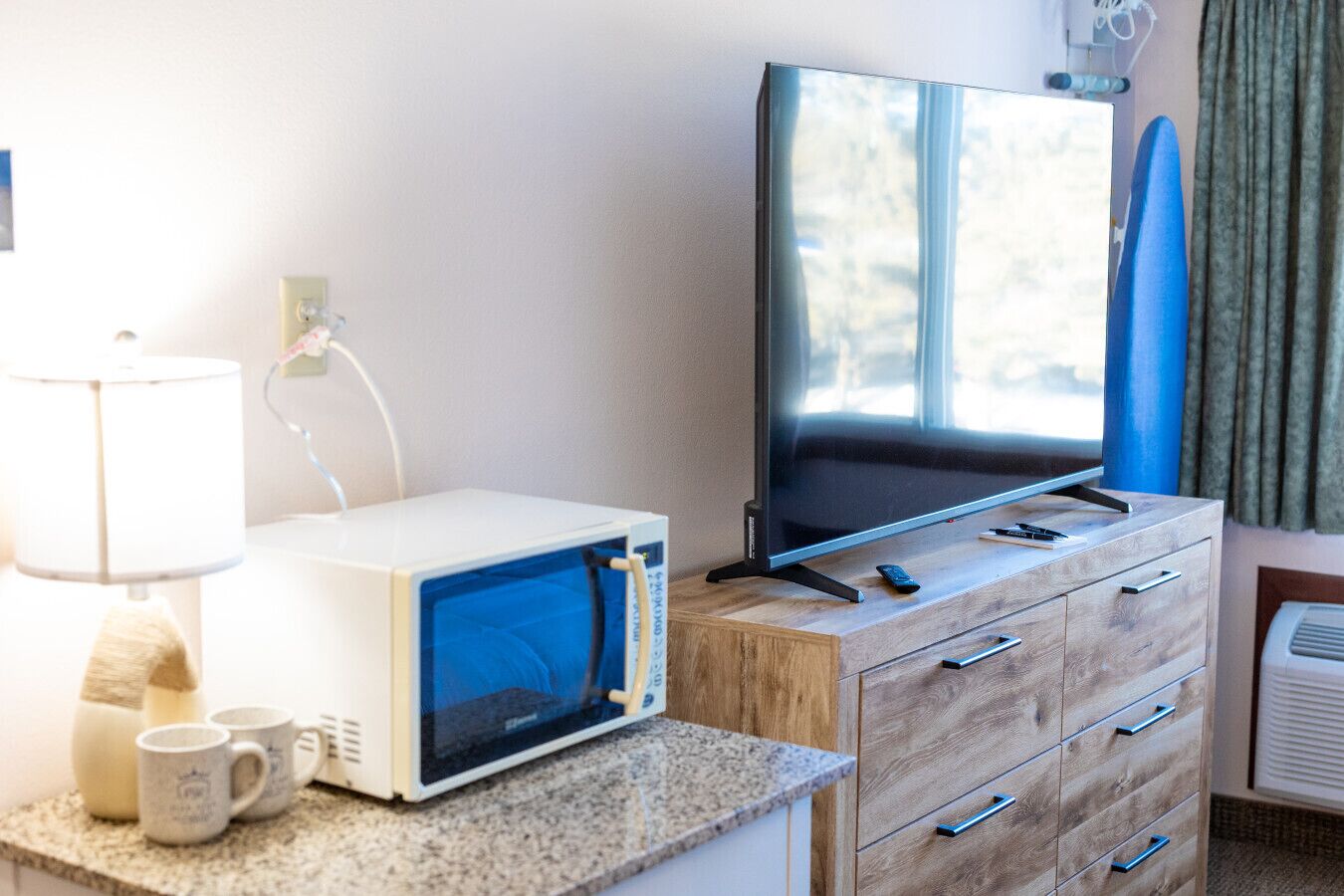 A room with a television on a wooden dresser, a microwave on a counter, and a lamp.