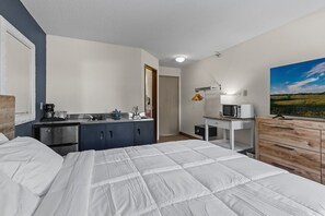 A hotel room with a bed, a kitchenette, a desk with a computer, and a painting on the wall.