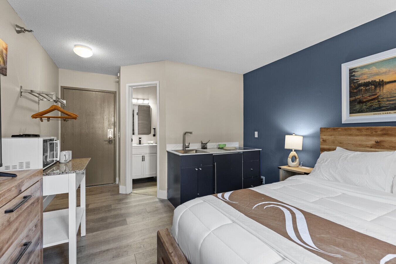 Une chambre d’hôtel comprenant un lit, un bureau avec un micro-ondes, une commode, un miroir, une table de chevet avec une lampe et un tableau au mur.