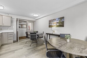 Eine moderne Küche mit weißen Schränken, ein Essbereich mit einem runden Holztisch und schwarzen Stühlen sowie ein Landschaftsbild an der Wand.