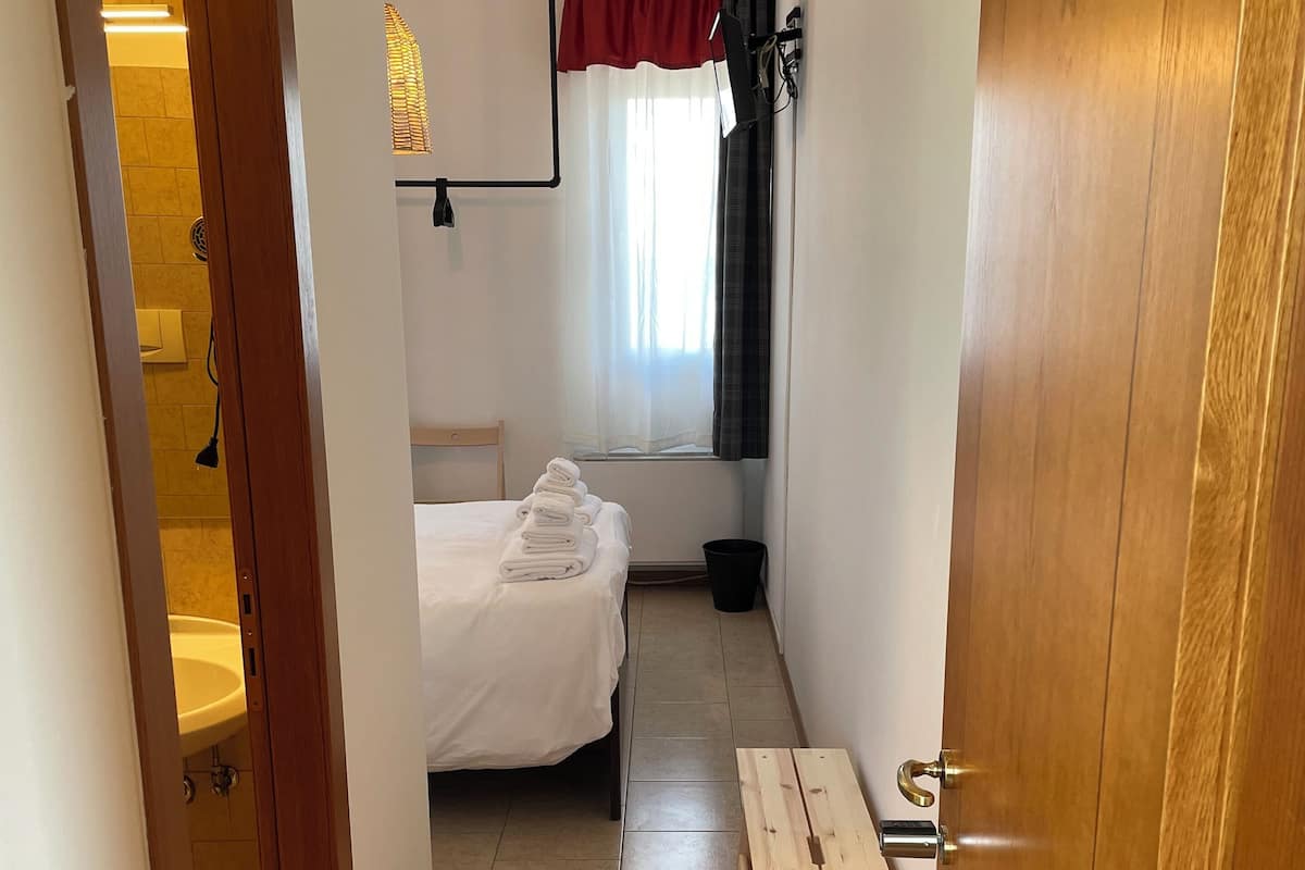 A hotel room with a bed, a wooden bench, a bathroom with a sink, and a window with curtains.