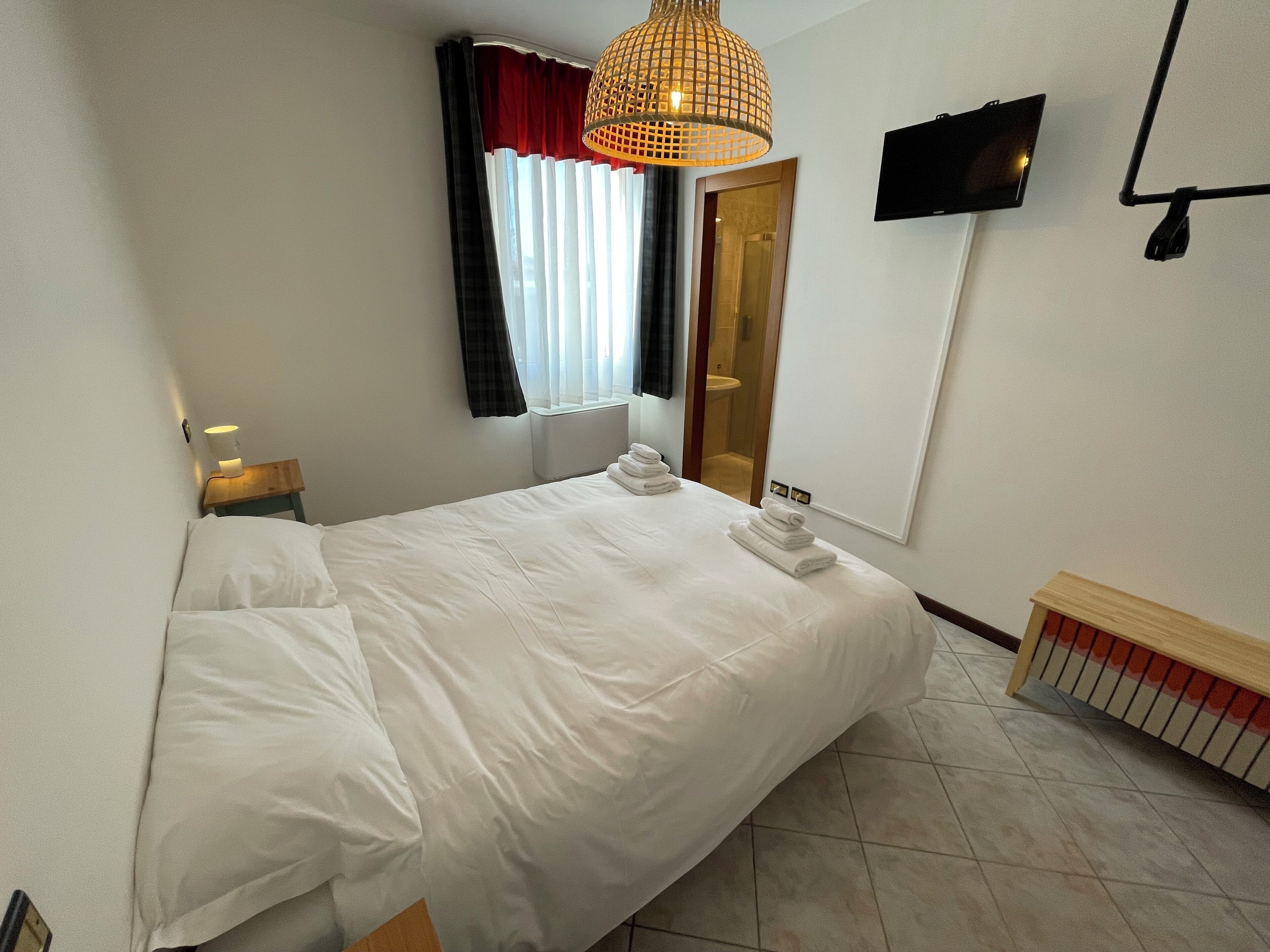 A hotel room with a large bed, a bedside table with a lamp, a TV mounted on the wall, and a window with curtains.