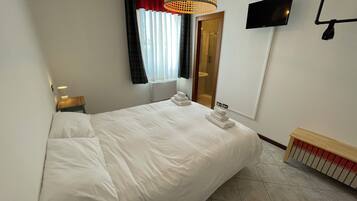 A hotel room with a large bed, a bedside table with a lamp, a TV mounted on the wall, and a window with curtains.