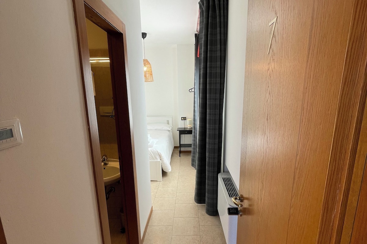 A narrow hallway with a wooden door on the right, a white wall, and a bed with a white headboard in the background.