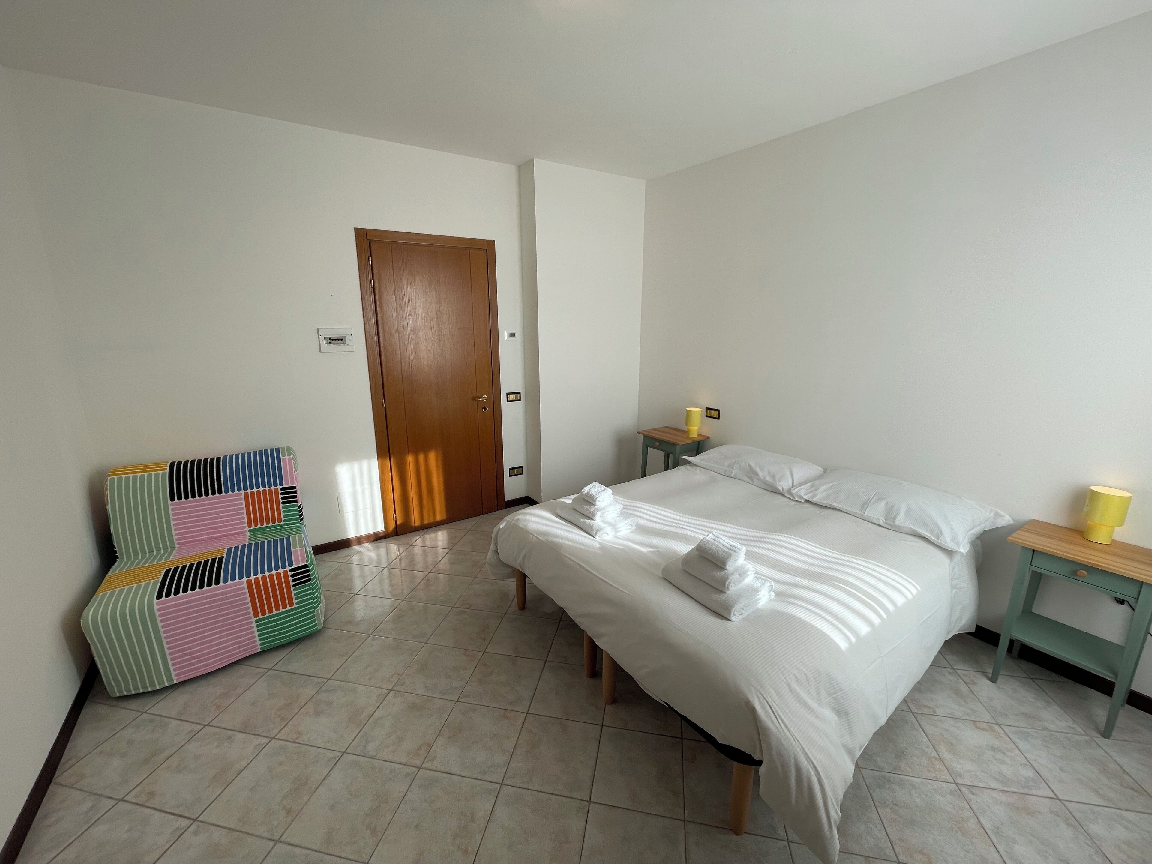 A bedroom with a bed, a bedside table with a lamp, a wardrobe, and a colorful bench.