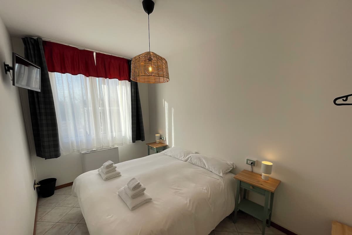 A hotel room with a large bed, a bedside table with a lamp, a TV mounted on the wall, and a window with curtains.