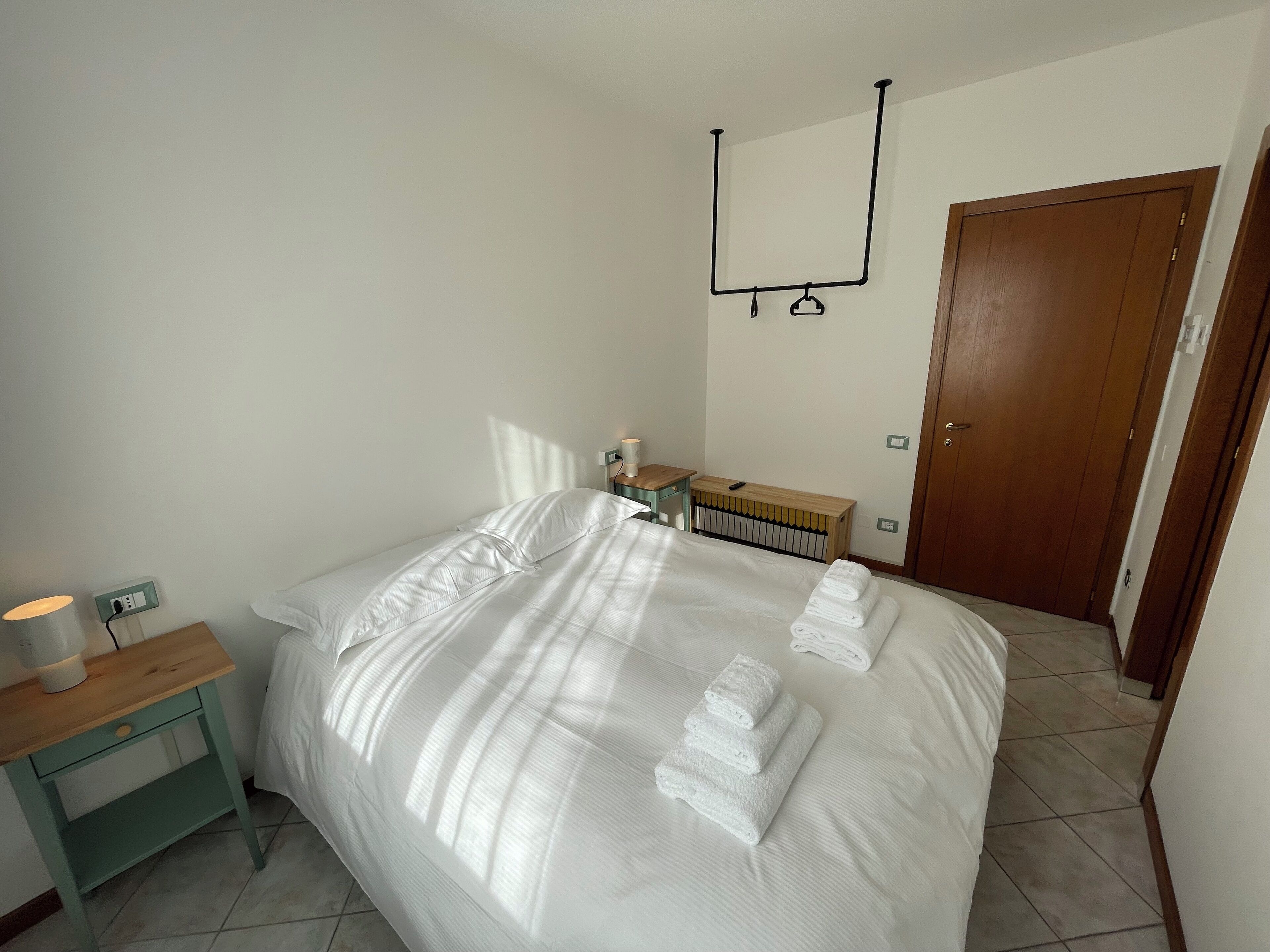 Una cama bien tendida con ropa de cama y toallas blancas, una mesita de noche con lámpara, una mesita auxiliar de madera y un radiador.