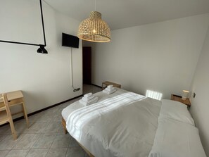 A neatly made bed with white linens, a wooden nightstand, a small wooden table, a woven pendant light, and a flat-screen TV mounted on the wall. - Albergaria (Rossano Veneto)