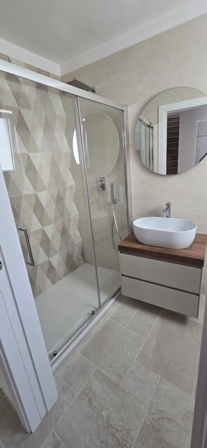 Une salle de bain moderne avec une douche à l’italienne en verre, un lavabo blanc posé sur un plan de travail en bois et un grand miroir au-dessus du lavabo.