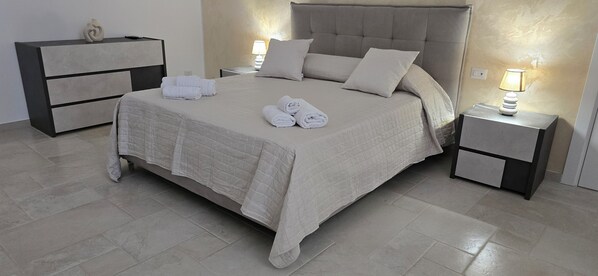 Un lit bien fait, recouvert d’une courtepointe matelassée, avec deux oreillers et deux serviettes pliées posées dessus. Il y a deux tables de chevet, chacune avec une lampe. Une commode avec un miroir est adossée au mur.