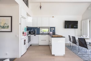 Une cuisine moderne avec des meubles blancs, un îlot central, un coin repas avec des chaises et un téléviseur à écran plat fixé au mur.