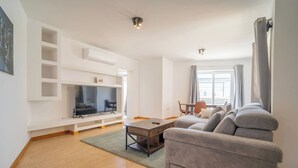 A modern living room with a flat-screen TV, a grey sofa, a wooden coffee table, and a dining area with a table and chairs.