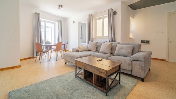 A modern living room with a grey sofa, wooden coffee table, and a dining area with chairs and a table.