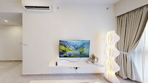 A modern living room with a flat-screen TV mounted on a white wall, a decorative lamp, and a large window with curtains. - SOHO Suites KLCC by noon (Kuala Lumpur)