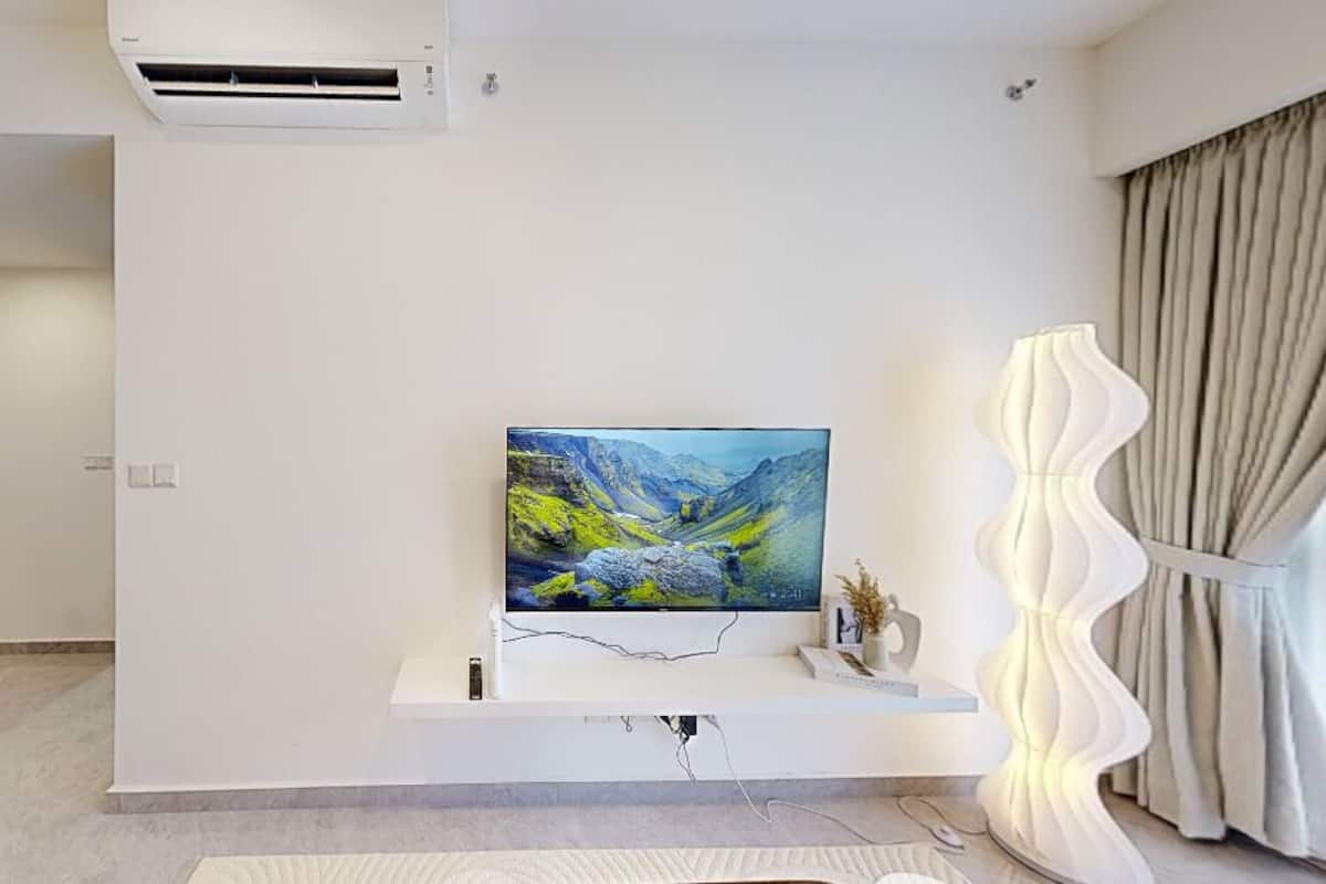A modern living room with a flat-screen TV mounted on a white wall, a decorative lamp, and a large window with curtains.