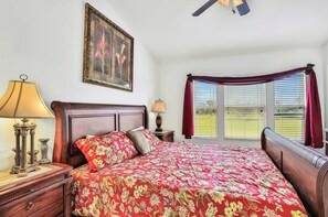 A bedroom with a wooden headboard, a floral bedspread, a nightstand with a lamp, a framed picture on the wall, and a window with blinds.