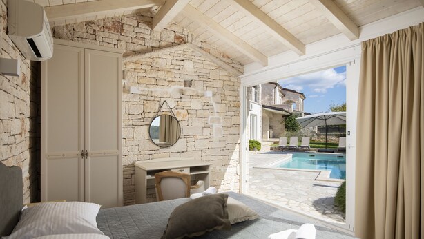 A bedroom with a stone wall, a bed with pillows, a desk, and a view of a pool and outdoor area. - Villa Mason with Heated Pool and mini Wellness (Rovinjsko Selo)