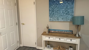 Un couloir avec une porte blanche, un tableau représentant l’océan accroché au mur, une console en bois surmontée d’une lampe, et divers objets décoratifs.