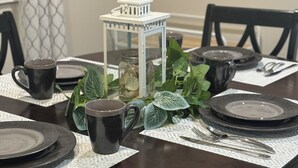 Une table de salle à manger ronde est dressée avec des assiettes noires, des tasses sombres et des couverts sur des sets de table blancs. Une lanterne blanche avec un chandelier est placée au centre, entourée de feuilles vertes.