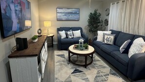 Un salon avec un canapé bleu foncé, une table basse, un téléviseur fixé au mur, un ventilateur de plafond et un tapis à motifs géométriques.