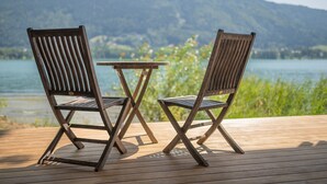 Dos sillas de madera y una mesita en una terraza de madera, con un lago y montañas al fondo.