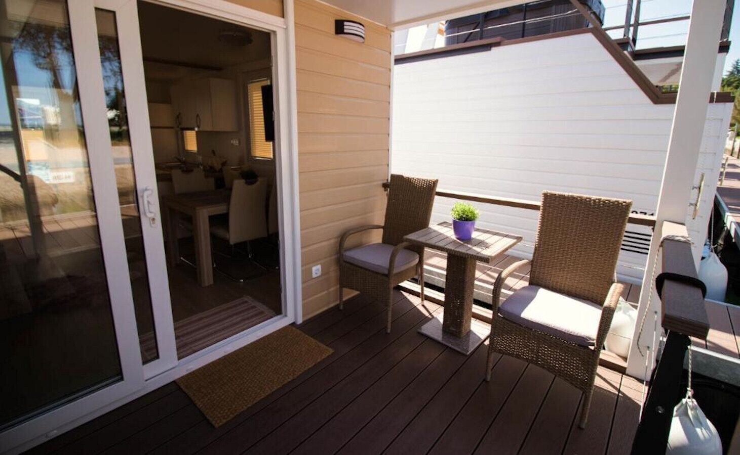 An outdoor deck with a sliding glass door leading to a kitchen, wicker chairs, and a small table with a potted plant.