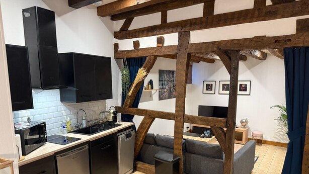 A modern kitchen with black cabinets, a microwave, and a dishwasher. There is a wooden beam structure in the center, and a gray sofa in the living area. - Charming Solognese house for 4 people (Pierrefitte-sur-Sauldre)